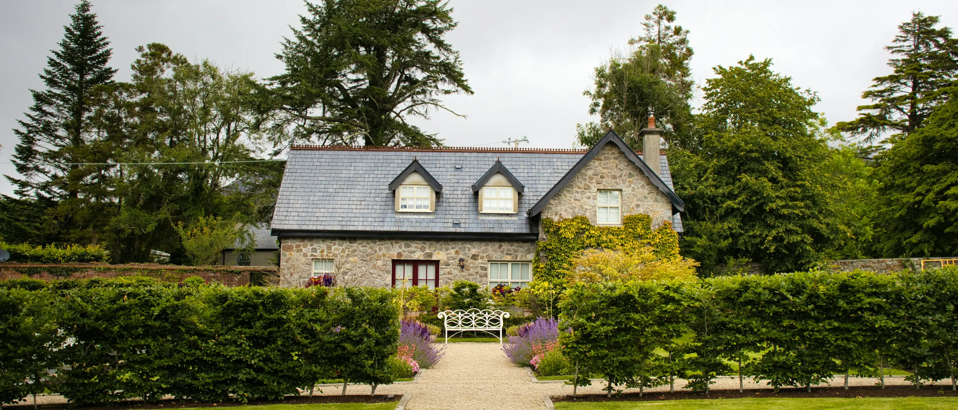 Ein steinernes Landhaus hinter geschnittenen Hecken, davor ein gepflegter Garten mit Weg zur Haustür und alte Bäume.